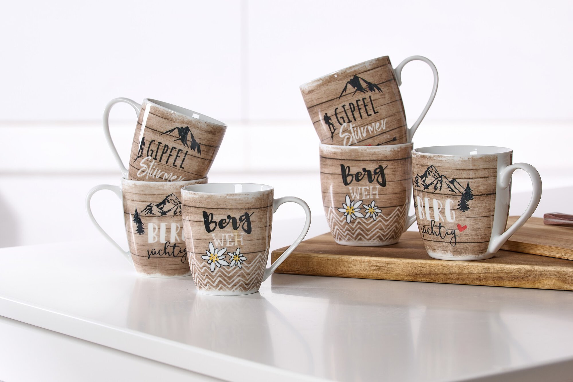 Sechs Kaffeetassen mit Berg-Motiven und Schriftzügen auf wei